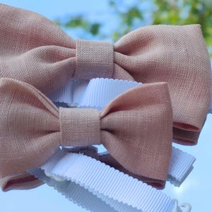 Peut inclure: Deux nœuds papillon rose poudré en tissu texturé, chacun doté d'un nœud central et d'une sangle réglable en ruban gros-grain blanc avec une fermeture à crochet en plastique. Les nœuds papillon sont superposés, le plus grand au-dessus du petit.