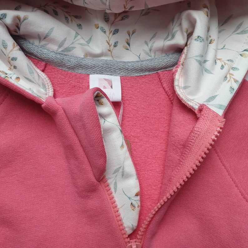 Peut inclure: Un sweat &agrave; capuche rose avec une doublure &agrave; imprim&eacute; floral blanc. La fermeture &eacute;clair est visible et le tissu est doux et confortable.