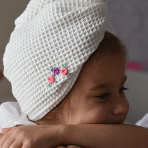 May include: A white waffle weave turban with pink and purple embroidered flowers. The turban is worn by a young girl with brown hair.
