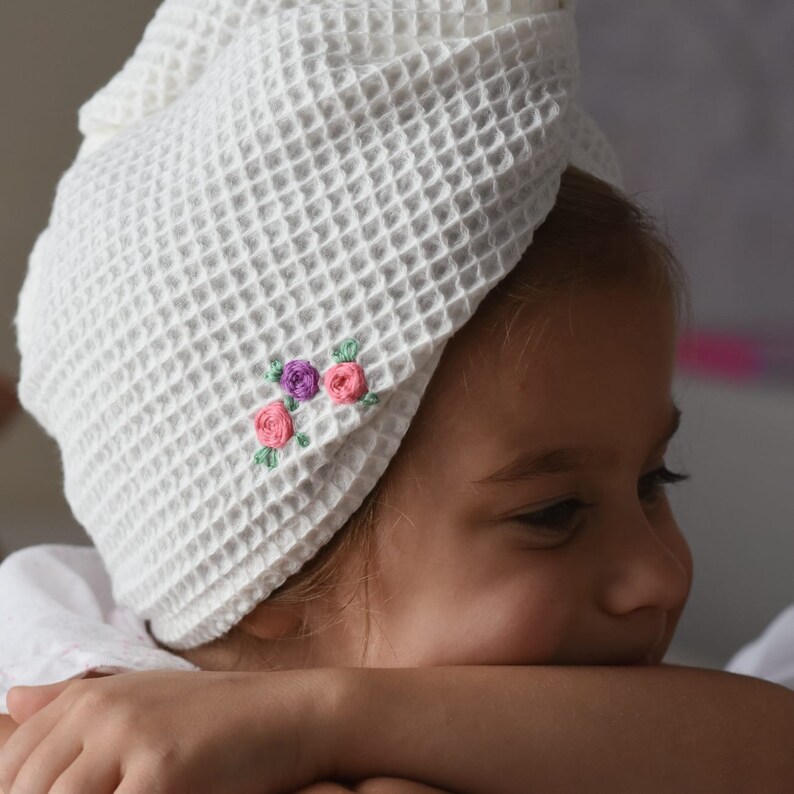 May include: A white waffle weave turban with pink and purple embroidered flowers. The turban is worn by a young girl with brown hair.