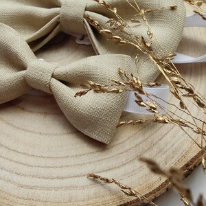 Puede incluir: Una pajarita de lino beige con una correa ajustable blanca, expuesta sobre una rodaja de madera con trigo dorado seco.