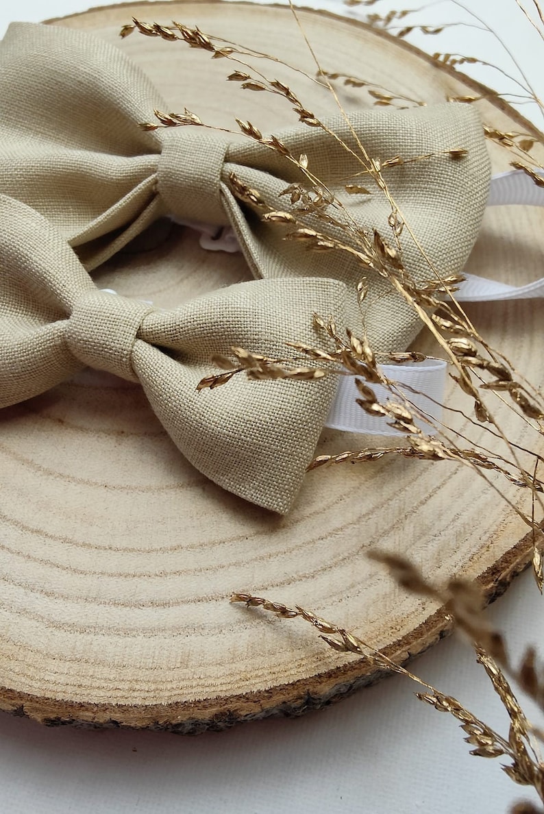 Puede incluir: Una pajarita de lino beige con una correa ajustable blanca, expuesta sobre una rodaja de madera con trigo dorado seco.