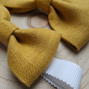 Op de afbeelding: Close-up van een mosterdgele vlinderdas van textiel. Een wit lint met geschulpte rand ligt naast de vlinderdas. De vlinderdas is een klassiek accessoire.