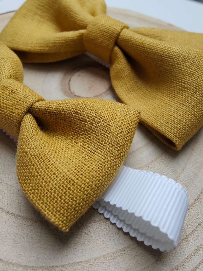 Op de afbeelding: Close-up van een mosterdgele vlinderdas van textiel. Een wit lint met geschulpte rand ligt naast de vlinderdas. De vlinderdas is een klassiek accessoire.