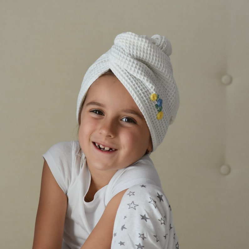 May include: A young girl wearing a white waffle weave turban with embroidered flowers on the side. She is wearing a white shirt with grey stars on the sleeve.