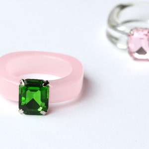May include: Two rings are displayed against a white background. One is a light pink, square-cut ring with a large, emerald-green gemstone. The other ring is clear with a pink gemstone. Both rings have a modern, minimalist design.
