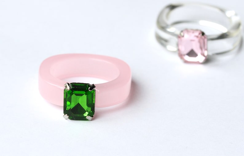 May include: Two rings are displayed against a white background. One is a light pink, square-cut ring with a large, emerald-green gemstone. The other ring is clear with a pink gemstone. Both rings have a modern, minimalist design.