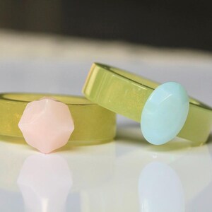 May include: Two rings with a light green band and a gemstone accent. One ring has a pink gemstone, and the other has a light blue gemstone. The rings are on a white surface.