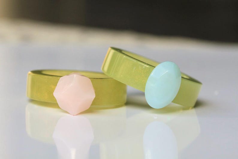 May include: Two rings with a light green band and a gemstone accent. One ring has a pink gemstone, and the other has a light blue gemstone. The rings are on a white surface.