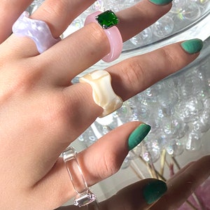 May include: A collection of colorful resin rings. The rings are in various shapes and colors, including light purple, pink, clear, and off-white. One ring features a square green gemstone. The rings are displayed on a reflective surface.