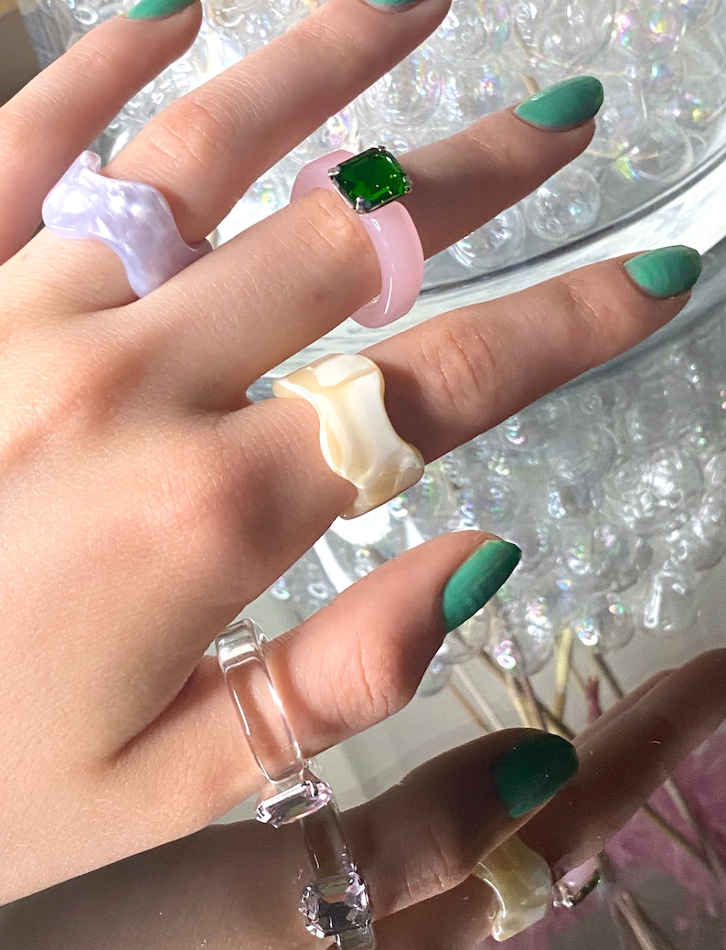 May include: A collection of colorful resin rings. The rings are in various shapes and colors, including light purple, pink, clear, and off-white. One ring features a square green gemstone. The rings are displayed on a reflective surface.