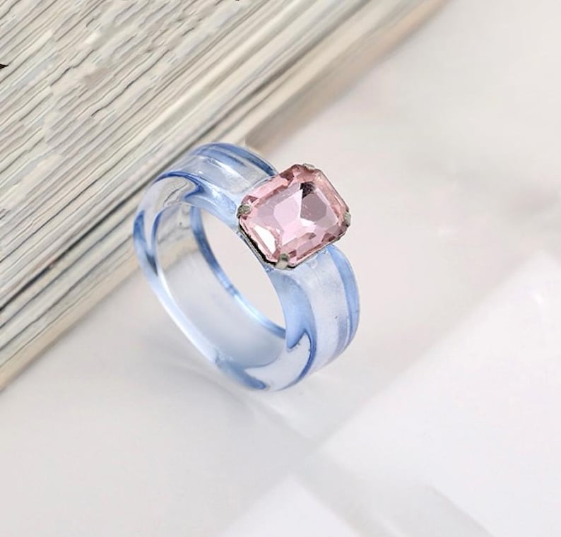 May include: A clear, light blue ring with a square, pink gemstone. The ring has a transparent band with a subtle blue hue, and the gemstone is set in a silver-colored metal. The ring is a fashion accessory.