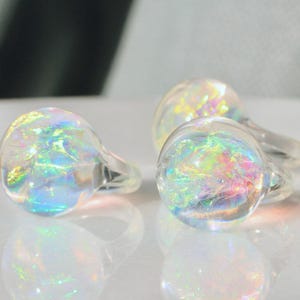 May include: Three clear glass rings with iridescent, rainbow-coloured inclusions. The rings are round with a small, clear band. The rings are on a white surface, reflecting the light and colours.