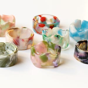 May include: Eight resin rings in various colours. Each ring has a unique marbled pattern in shades of pink, green, blue, and purple. The rings have a chunky, rounded design, ideal for adding a pop of colour to any outfit.