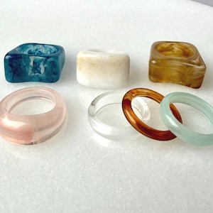 Peut inclure: Plusieurs bagues en résine colorées de différentes formes et tailles. Les bagues comprennent une bague carrée bleue, une bague carrée ambre, une bague rose, une bague transparente, une bague marron et une bague vert clair.