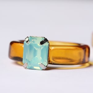 May include: A close-up of a ring with a rectangular, light blue gemstone set in a silver metal. The ring band is a transparent amber color. Another ring is slightly visible in the background.