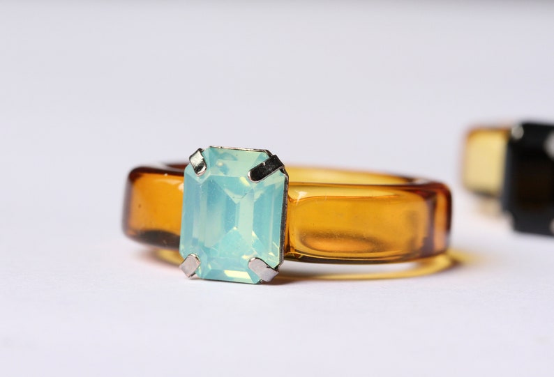 May include: A close-up of a ring with a rectangular, light blue gemstone set in a silver metal. The ring band is a transparent amber color. Another ring is slightly visible in the background.