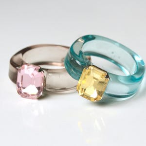 May include: Two chunky rings with large, rectangular gemstones. One ring is clear with a pink gem, the other is a translucent teal with a yellow gem. The rings are made of a clear, glossy material.