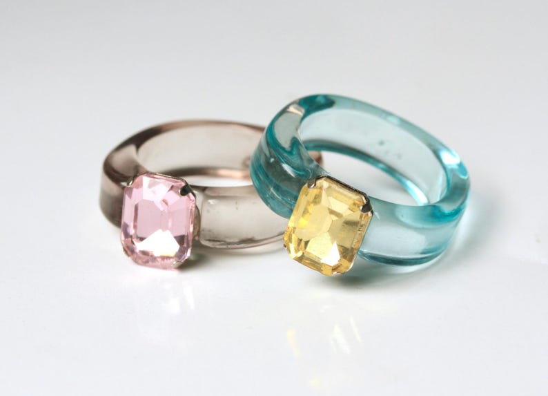May include: Two chunky rings with large, rectangular gemstones. One ring is clear with a pink gem, the other is a translucent teal with a yellow gem. The rings are made of a clear, glossy material.