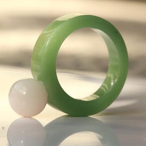 May include: A light green jade ring with a smooth, polished surface. A small, round, white stone rests next to the ring. The ring and stone are reflected on a shiny surface.