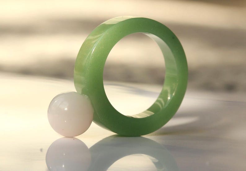 May include: A light green jade ring with a smooth, polished surface. A small, round, white stone rests next to the ring. The ring and stone are reflected on a shiny surface.
