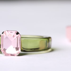 May include: A close-up of a ring with a light pink rectangular gemstone set on a transparent olive green band. Another ring with a pink and green gemstone is slightly out of focus in the background. The rings are on a white surface.