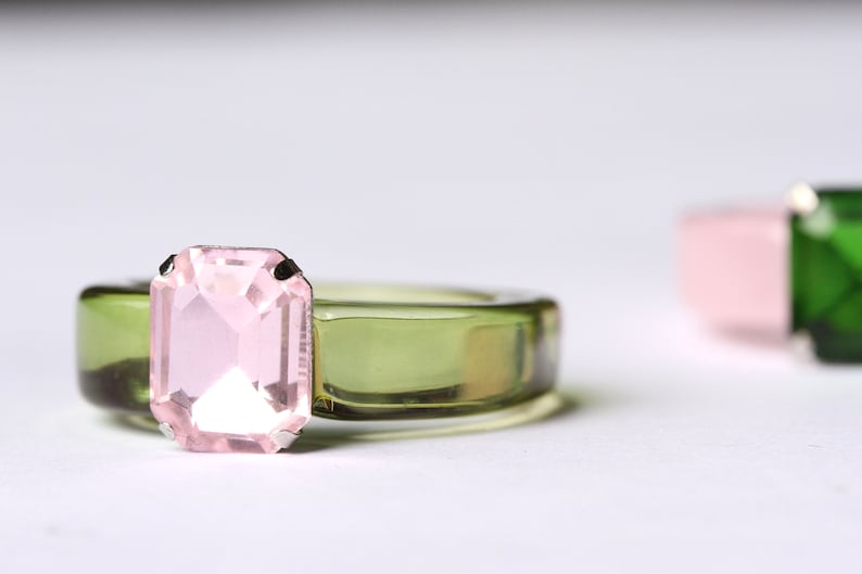 May include: A close-up of a ring with a light pink rectangular gemstone set on a transparent olive green band. Another ring with a pink and green gemstone is slightly out of focus in the background. The rings are on a white surface.
