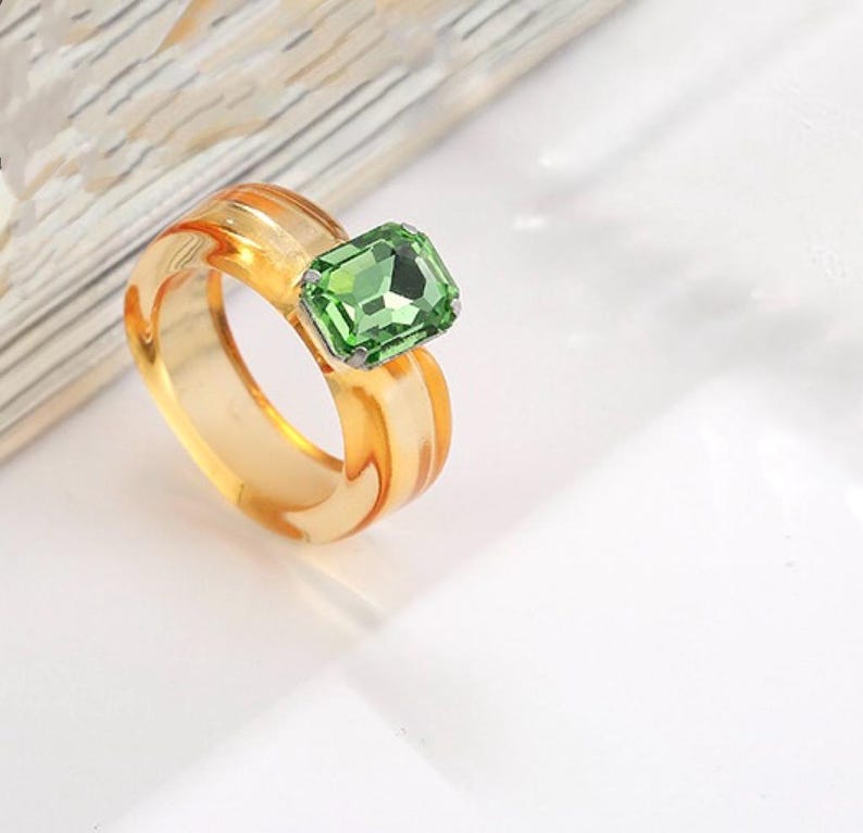 May include: A clear, amber-colored ring with a large, emerald-cut green gemstone. The ring has a thick band with a slightly textured design. The gemstone is set in a silver-colored metal.