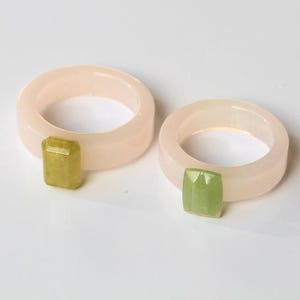 May include: Two rings with a pale pink band and a rectangular, faceted gemstone. One gemstone is yellow-green, the other is green. The rings are on a white surface.