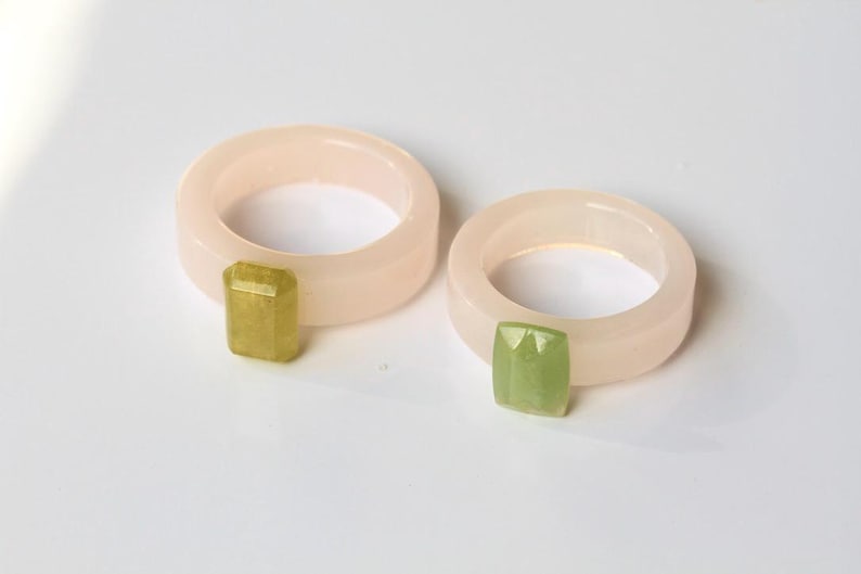 May include: Two rings with a pale pink band and a rectangular, faceted gemstone. One gemstone is yellow-green, the other is green. The rings are on a white surface.