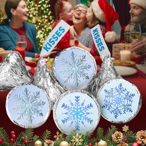 May include: A set of six silver foil-wrapped chocolate kisses with snowflake designs and "Kisses" labels. The kisses are arranged on a table with a Christmas tree and family in the background.