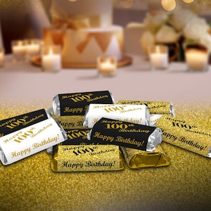 May include: A close-up of several small chocolate bars wrapped in gold foil with black and white labels. The labels have gold lettering that says "Happy 100th Birthday!" and "Happy Birthday!"