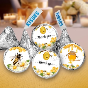 May include: Silver foil-wrapped chocolate candies with "Kisses" paper flags. The candy tops feature bee-themed designs with honeycombs, bees, and the words "Thank you". The candies are on a yellow surface.