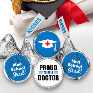 May include: A set of four round stickers for Hershey's Kisses chocolates. The stickers feature a blue background with white text and graphics. The stickers say "Med School Grad!" with a stethoscope, "Proud to be a Doctor" with three lines, and "Med School Grad!" with a graduation cap and cross. One sticker has a blue background with a white graduation cap and cross.