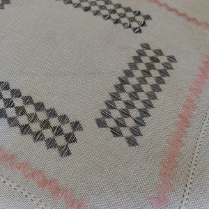 May include: A white tablecloth with a checkered pattern in black and a pink border. The tablecloth has a square shape and is made of a soft, woven fabric.