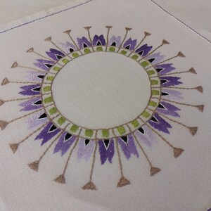 May include: A white linen napkin with an embroidered design of a purple and green circle with a white center.