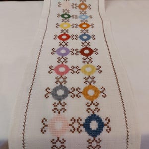 May include: White table runner with a brown embroidered border and a repeating pattern of colorful circles in a cross shape.