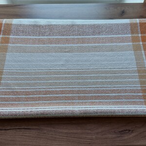 May include: A rectangular, woven table runner with a plaid and striped pattern. The runner features a color palette of brown, orange, and white. The design includes both horizontal stripes and vertical plaid sections, creating a textured appearance.