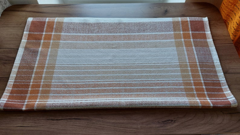 May include: A rectangular, woven table runner with a plaid and striped pattern. The runner features a color palette of brown, orange, and white. The design includes both horizontal stripes and vertical plaid sections, creating a textured appearance.