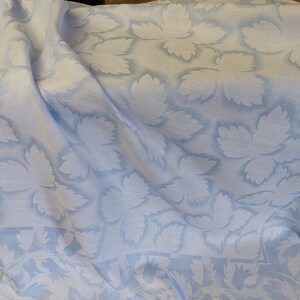 May include: A white linen tablecloth with a blue leaf pattern. The tablecloth is folded and draped over a surface.