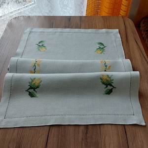 May include: A white linen table runner with embroidered yellow flowers and green leaves. The runner is folded in thirds and has a black stitched border.
