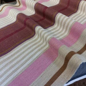 May include: Close-up of a woven textile with a striped pattern. The fabric features alternating stripes of beige, pink, and burgundy, with thin brown stripes. The textile is draped, showing the texture and weave.