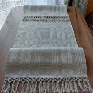 May include: A beige linen table runner with a checkered pattern and a crocheted fringe. The runner is folded in half and laid on a wooden surface.