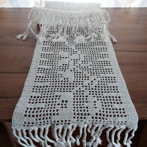 May include: White crocheted table runner with a delicate floral pattern and fringe trim.