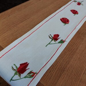 May include: White linen table runner with a red embroidered border and red rose designs along the center.