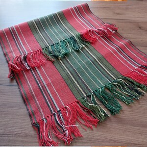 May include: A red and green striped table runner with fringe on the ends. The runner is made of woven fabric and has a rustic look.