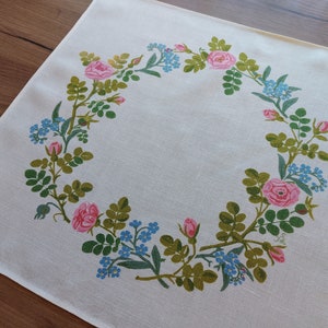 May include: White tablecloth with a floral design featuring pink roses and blue forget-me-nots arranged in a wreath pattern.