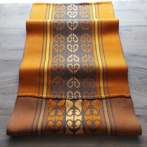 Op de afbeelding: Een geweven tafelloper in oranje- en bruintinten met een herhalend hartvormig patroon in het midden. De loper heeft een rand van oranje en gele strepen. De stof is opgevouwen, waardoor de textuur en het ontwerp zichtbaar zijn.