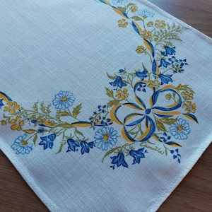 May include: White linen table runner with a blue and yellow floral design. The design features a large bow with blue and yellow ribbons, surrounded by bluebells and daisies. The text "Camilla" is printed in the lower right corner.