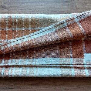 May include: Folded plaid kitchen towel in shades of brown, orange, and cream. The woven fabric features a classic check pattern. A small, blank white tag is sewn on the towel. The towel is folded neatly.